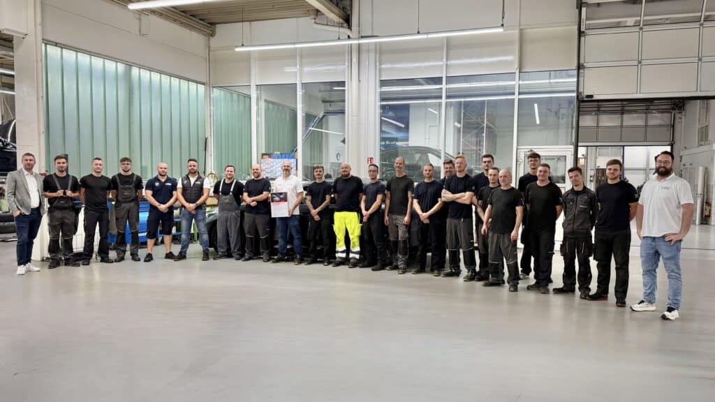 Großes Werkstatt-Team posiert gemeinsam in heller Autohaus-Halle, eine Person hält eine Urkunde.
